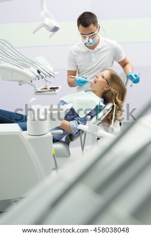 Dental cabinet with a male dentist and a female patient. He is in the white uniform with blue latex gloves, blue mask, binocular loupes. She is in the blue shirt and jeans on the patient chair. She