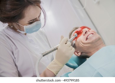 Dental Care Concept Stomatology Inspection. The Patient Lies In A Chair In Dentistry In Front Of Him A Hand With Drill. The Dentist Is In The Office Beside The Chair With The Patient