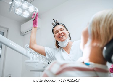 Dental clinic patient appointment in modern medical ward. Dentist doctor sincerely smiling, pointing dental light at young Female ready for teeth surgery. Health care and medicare industry image. - Powered by Shutterstock