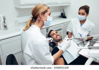 Dental Doctor Looking At A Display Monitor While Treating A Male Patient. Female Dentist Wearing Face Mask With Male Patient Sitting In Dentist's Chair And Her Assistant Sitting By In Dental Clinic.