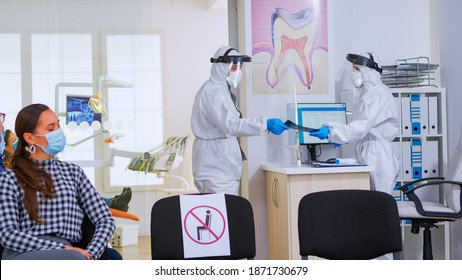 Dental Doctor Wearing Ppe Suit Against Covid-19 Holding Teeth X-ray While Woman With Mask Waiting In Reception Respecting Social Distance. Concept Of New Normal Dentist Visit In Coronavirus Outbreak.