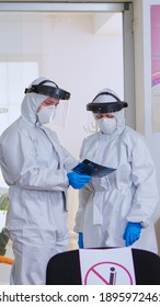 Dental Doctors With Protection Suit Analysing Teeth X-ray In Reception While Patient Waiting On Chair, Respecting Social Distance. Concept Of New Normal Dentist Visit In Coronavirus Outbreak.