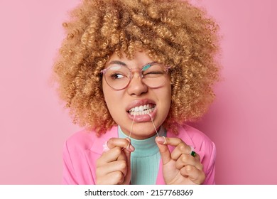 Dental health care concept. Beautiful young woman with curly bushy hair cleans teeth with dental floss wears transparent eyesglasses isolated over pink background shows how to floss correctly - Powered by Shutterstock