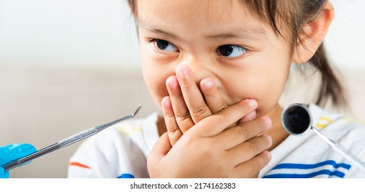 Dental kid examination. Doctor examines oral cavity of child uses mouth mirror to check teeth cavity but little girl cover mouth no need to checking she afraid, scared and closes his mouth with hands - Powered by Shutterstock