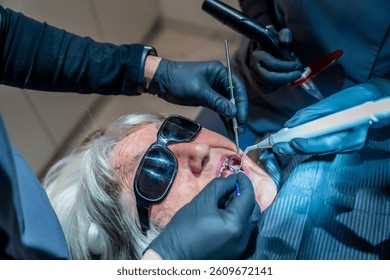 Dental professionals using specialized tools are performing a teeth whitening procedure on a senior woman patient wearing protective eyewear