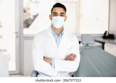 Dental Services. Portrait Of Handsome Arab Dentist Doctor In Uniform And Protective Medical Face Mask Posing At Workplace In Modern Clinic Interior, Standing With Folded Arms, Ready For Check Up - Powered by Shutterstock