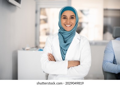 Dental Services. Portrait Of Smiling Islamic Dentist Woman Standing With Folded Arms At Workplace In Stomatologic Clinic, Happy Muslim Stomatologist Lady In Hijab Looking At Camera, Closeup