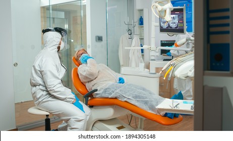 Dental Specialist With Coverall Talking With Senior Patient Before Examination During Pandemic Covid-19. Concept Of New Normal Dentist Visit In Coronavirus Outbreak Wearing Protective Suit And Face