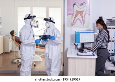 Dentist Assistant Discussing With Doctor Patient Diagnosis Keeping Social Distancing Dressed In Ppe Suit Face Shiled, During Global Pandemic With Coronavirus Holding X-ray.