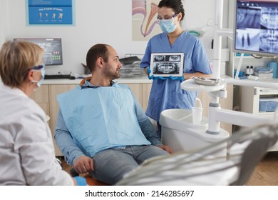 Dentist Assistant With Face Mask Holding Tabet Computer Explaining Teeth Radiography To Sick Patient During Dentistry Consultation In Stomatological Office Room. Team Working At Caries Treatment