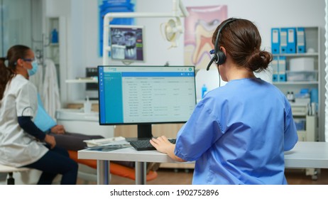 Dentist assistant making appointments using headset sitting in front on computer while doctor is working with patient in background examining teeth problem. Nurse taking notes in stomatological office - Powered by Shutterstock