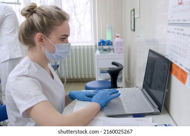 Dentist Assistant Working On Laptop In Dental Office Wearing Medical Gloves And Mask. Young Doctor In White Uniform Sit At Desk In Clinic With Computer. Healthcare, Medicine, Dentistry And Technology