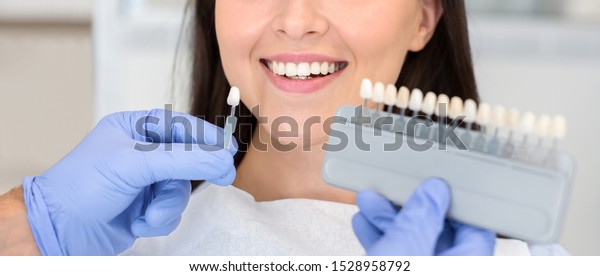 Dentist in blue medical\
gloves applying sample from tooth enamel scale to happy woman\
patient teeth to pick up right shade, teeth bleaching procedure,\
cropped, panorama