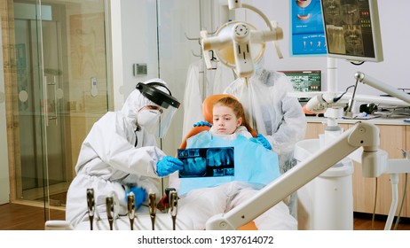 Dentist With Coverall Holding Mouth X-ray Image Child Patient Talking With Patient Mother During Global Pandemic. Assistant And Doctor Talking Wearing Suit, Coverall, Protection Suit, Mask, Gloves