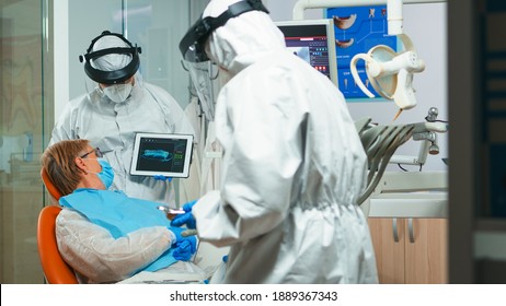 Dentista en forma encubierta usando un comprimido que explique rayos x dentales al paciente en el consultorio estomatológico durante el coronavirus. Hombre con el escudo de la cara, cubierto, mostrando máscara a la mujer radiografía usando dispositivo digital