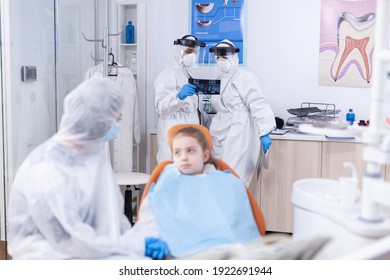 Dentist Doctor And Assistant Holding Dental Radiography Of Little Kid Patient In The Course Of Coronavirus. Stomatologist During Covid19 Wearing Ppe Suit Doing Teeth Procedure Of Child Sitting On