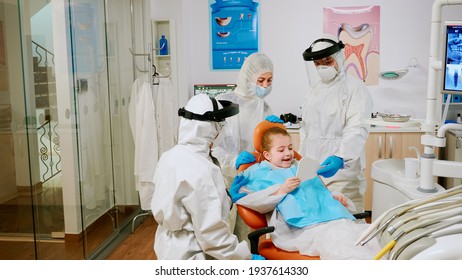 Dentist Doctor In Coverall Finishing Dental Treatment Of Child In Stomatological Clinic During Global Pandemic. Medical Team Wearing Protection Suit, Face Shield Mask And Gloves Examining Patient