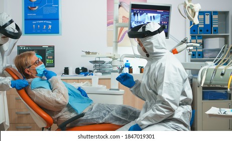 Dentist Doctor With Protection Equipment Talking With Old Patient Before Consultation During Covid-19 Epidemic. Concept Of New Normal Dentist Visit In Coronavirus Outbreak Wearing Protective Suit And