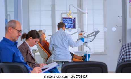 Dentist Doctor Working In Crowded Professional Dental Office With Senior Patient. Orthodontist Speaking To Woman Sitting On Stomatological Chair While People Waiting In Reception Area Filling In Forms