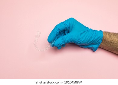 Dentist Doctor's Hand Wearing Blue Latex Gloves Holds A Clear Dental Aligner On Pink Background. Braces Dental Concept