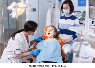 Dentist Doing Teeth Check Up Of Little Girl Sitting On Dental Chair Wearing Bib. Dentistry Specialist During Child Cavity Consultation In Stomatology Office Using Modern Technology.