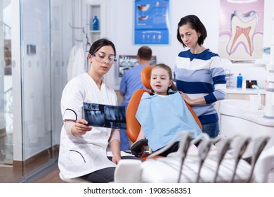 Dentist Examining Little Kid Radiography Sitting On Chair Wearing Dental Bib. Stomatologist Explaining Teeth Diagnosis To Mother Of Child In Health Clinic Holding X-ray.