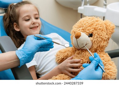 Dentist examining the teeth of a teddy bear on the hands of a little girl sitting in dentist chair. Girl playing as her teddy bear is patient in dentist office - Powered by Shutterstock