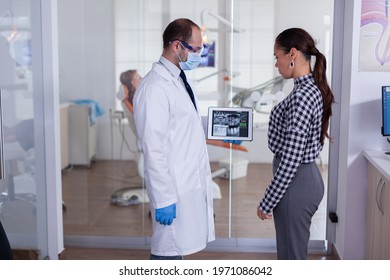 Dentist With Face Mask Showing Patient Dental X-ray In Stomatology Specialist Reception. Explaining Diagnosis During Appointment, Senior Man Sitting In Dental Chair.