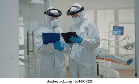 Dentist Giving Tablet To Dental Nurse Before Consultation Appointment With Patient. Stomatology Doctors Wearing Ppe Suits During Pandemic While Treating Man For Oral Healthcare