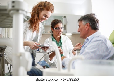 Dentist And Her Female Assistant In Dental Office Talking With Senior Patient And Preparing For Treatment.