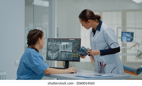 Dentist holding dentition x ray scan comparing to radiography on computer monitor at dental clinic. Nurse sitting at desk looking at screen for teeth examination and consultation