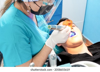 Dentist Latina Woman Dressed In Blue Attending To Latina Man Dressed In Black, With Ultraviolet Light Lamp Drying A Resin Or Amalgam In Patient's Mouth, Dental Office