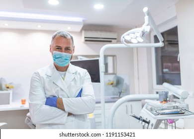 Dentist In Mask During The Dental Surgery. Smiling Dentist Leaning Against Dentists Chair In Dental Clinic. Man Dentisit In Office. Portrait Of Male Dentist .He Standing In His Dentist Office.
