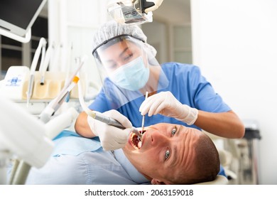 Dentist In Mask And Face Shield Treating Male Patient Using Dental Drill In Dentist Office