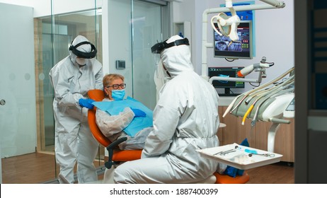 Dentist Nurse In Coverall Putting Dental Bib To Old Woman Before Stomatological Examination During Covid-19 Pandemic. Concept Of New Normal Dentist Visit In Coronavirus Outbreak Wearing Protective