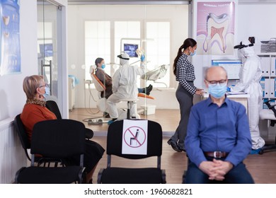 Dentist Nurse Dressed In Ppe Suit With Face Shiled Discussing With Patient In Stomatology Waiting Room. People Keeping Social Distancing As Prevention During Coronavirus Outbreak.