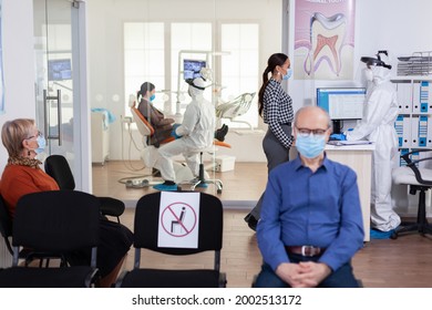 Dentist Nurse Dressed In Ppe Suit With Face Shiled Discussing With Patient In Stomatology Waiting Room. People Keeping Social Distancing As Prevention During Coronavirus Outbreak.