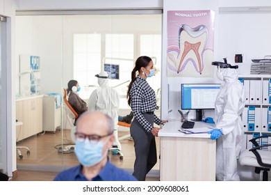 Dentist Nurse Dressed In Ppe Suit With Face Shiled Discussing With Patient In Stomatology Waiting Room. People Keeping Social Distancing As Prevention During Coronavirus Outbreak.