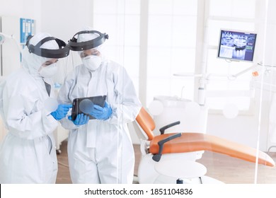 Dentist And Nurse During Covid-19 Outbreak In Ppe Suit Usint Tablet Pc. Stomatology Team In Dental Office Wearing Protective Suit Agasint Contagious Coronavirus During Global Pandemic.