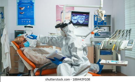 Dentist Nurse In Potective Suit Controlling Patient Temperature Before Dental Examination During Coronavirus Pandemic. Concept Of New Normal Dentist Visit In Coronavirus Outbreak Wearing Protective