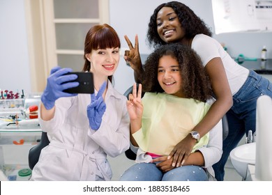 Dentist office, pediatric dentistry. Female pediatric dental specialist making selfie photo on smartphone, together with her patients, little curly mixed raced school girl and her young African mom.