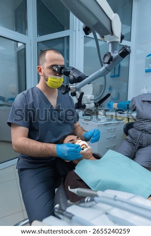 Dentist Performing a Procedure Under Microscope in Modern Dental Office with Patient Relaxed for Treatment and Assistant on Background Preparing Tools