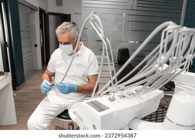 A dentist sits behind a dental chair and prepares tools for dental treatment. A senior, experienced male doctor works in a dental treatment and prosthetics center. - Powered by Shutterstock