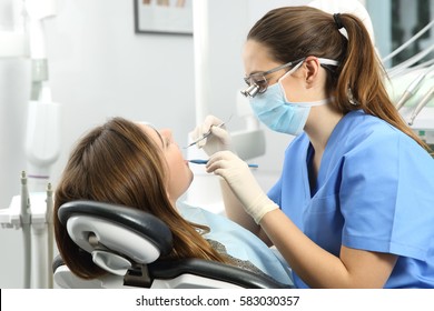 Dentist wearing eyeglasses gloves and mask examining a patient teeth with a dental probe and a mirror in a clinic box with equipment in the background - Powered by Shutterstock