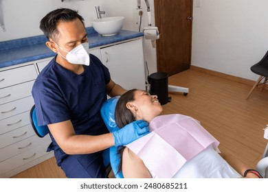 A dentist wearing a mask and blue gloves is gently adjusting the headrest for a relaxed patient in a dental chair in a modern dental clinic. The patient is smiling, feeling comfortable. - Powered by Shutterstock