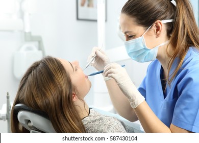 Dentist wearing mask gloves and blue uniform examining a patient teeth with a dental probe and a mirror in a box with medical equipment in the background - Powered by Shutterstock