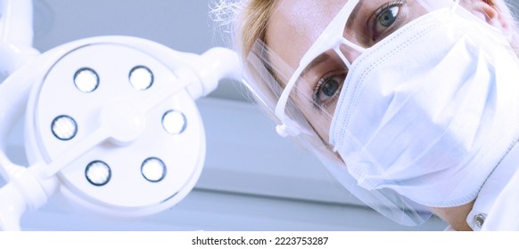 The Dentist, Wearing A Medical Mask And Goggles, Looks Straight Ahead. The Health Care Professional Examines The Patient With A Medical Light