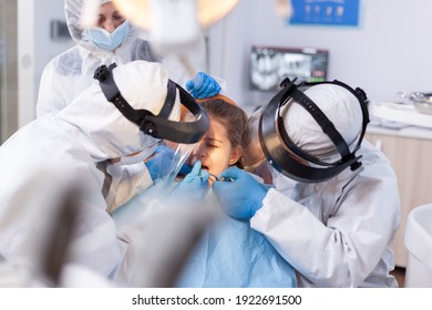 Dentist Woman Treating Little Girl Bad Tooth Using Drill With Help From Assistant Dressed In Coverall. Stomatology Team Wearing Ppe Suit During Covid19 Doing Procedure On Child Teeth.