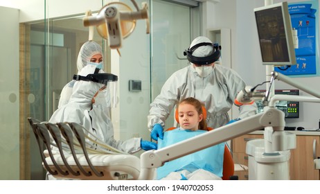 Dentistry Nurse In Coverall Putting Dental Bib To Child Before Stomatological Examination During Covid-19 Pandemic. Concept Of New Normal Dentist Visit In Coronavirus Outbreak Wearing Protective Suit