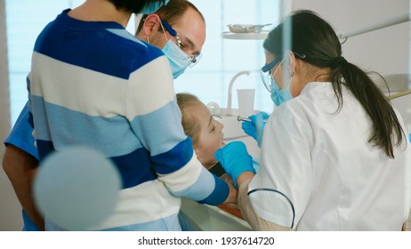 Dentists With Gloves And Mask Removes Tooth Decay Drilling Teeth During Surgery. Dentistry Doctors Treating Dental Problems Of Child, Mother Standing Near Stomatological Chair Talking With Girl.